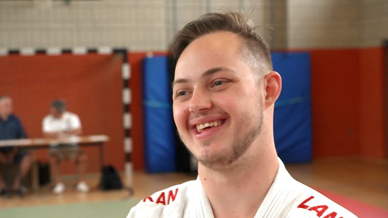 Ein junger Mann steht in einer Sporthalle und lächelt.